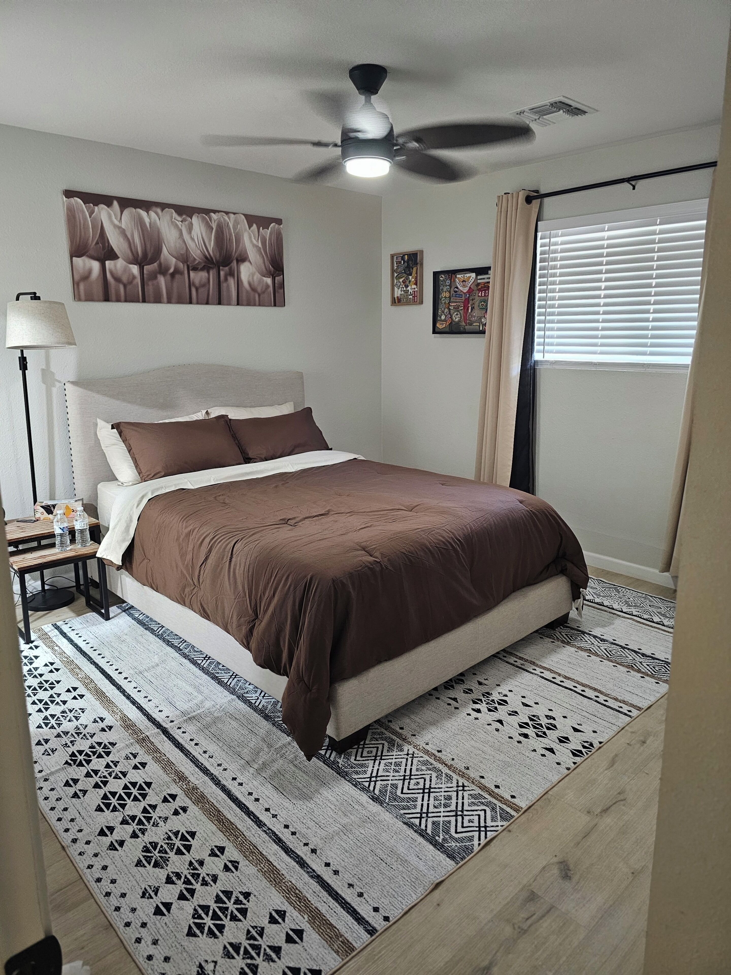 A bedroom with a bed, a ceiling fan, a nightstand, a lamp, and a window with blinds.