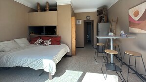 Een kleine kamer met een bed, een tafeltje met krukjes, een televisie en een koelkast.
