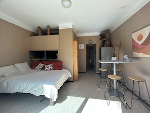 Ein kleines Zimmer mit einem Bett, einem Tisch mit Hocker, einem Fernseher und einem Kühlschrank.