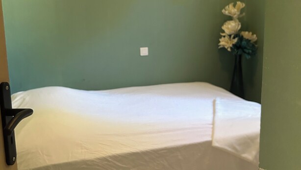 Een eenpersoonsbed met een witte matras en een plastic beschermhoes, een ingelijste afbeelding aan de muur en een vaas met bloemen aan de rechterkant.