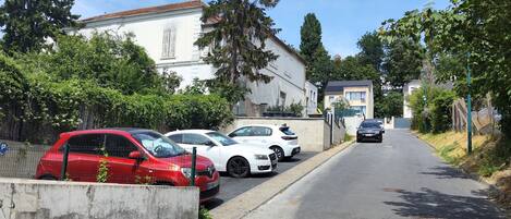 Une rue résidentielle bordée de voitures garées, avec un immeuble blanc et des arbres.