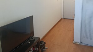 Ett rum med en platt-TV på ett stativ, ett trägolv, en vit dörr och en väggmonterad lampa.