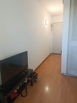 Ein Zimmer mit Flachbildfernseher auf einem Standfuß, Holzboden, weißer Tür und einer Wandleuchte.