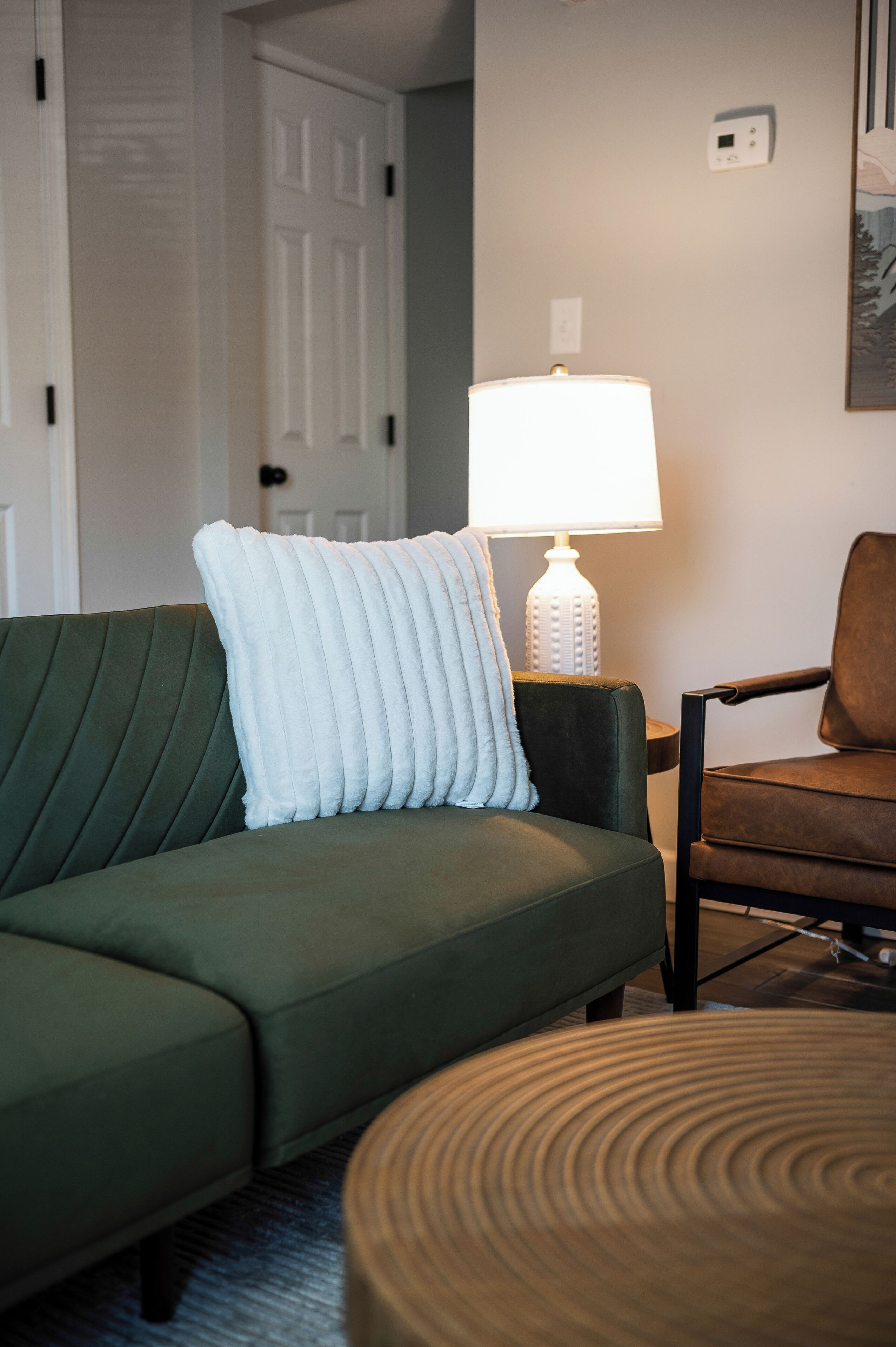 Un canapé vert avec un coussin blanc, une lampe blanche et une chaise marron dans une pièce dotée d’une porte et d’un thermostat fixé au mur.