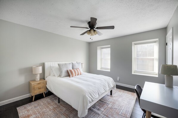 A bedroom with a bed, nightstand, lamp, ceiling fan, and two windows with blinds.