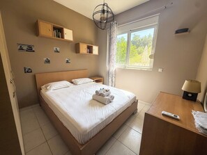 A hotel room with a large bed, bedside tables, a desk with a lamp, and a window with a view of greenery. - Villa Fountain (Pool, Garden, BBQ, Starlink, Wood House) (Afantou)