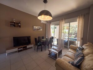 A living room with a sofa, a TV, a dining table with chairs, a ceiling light, and a view through the glass door. - Villa Fountain (Pool, Garden, BBQ, Starlink, Wood House) (Afantou)
