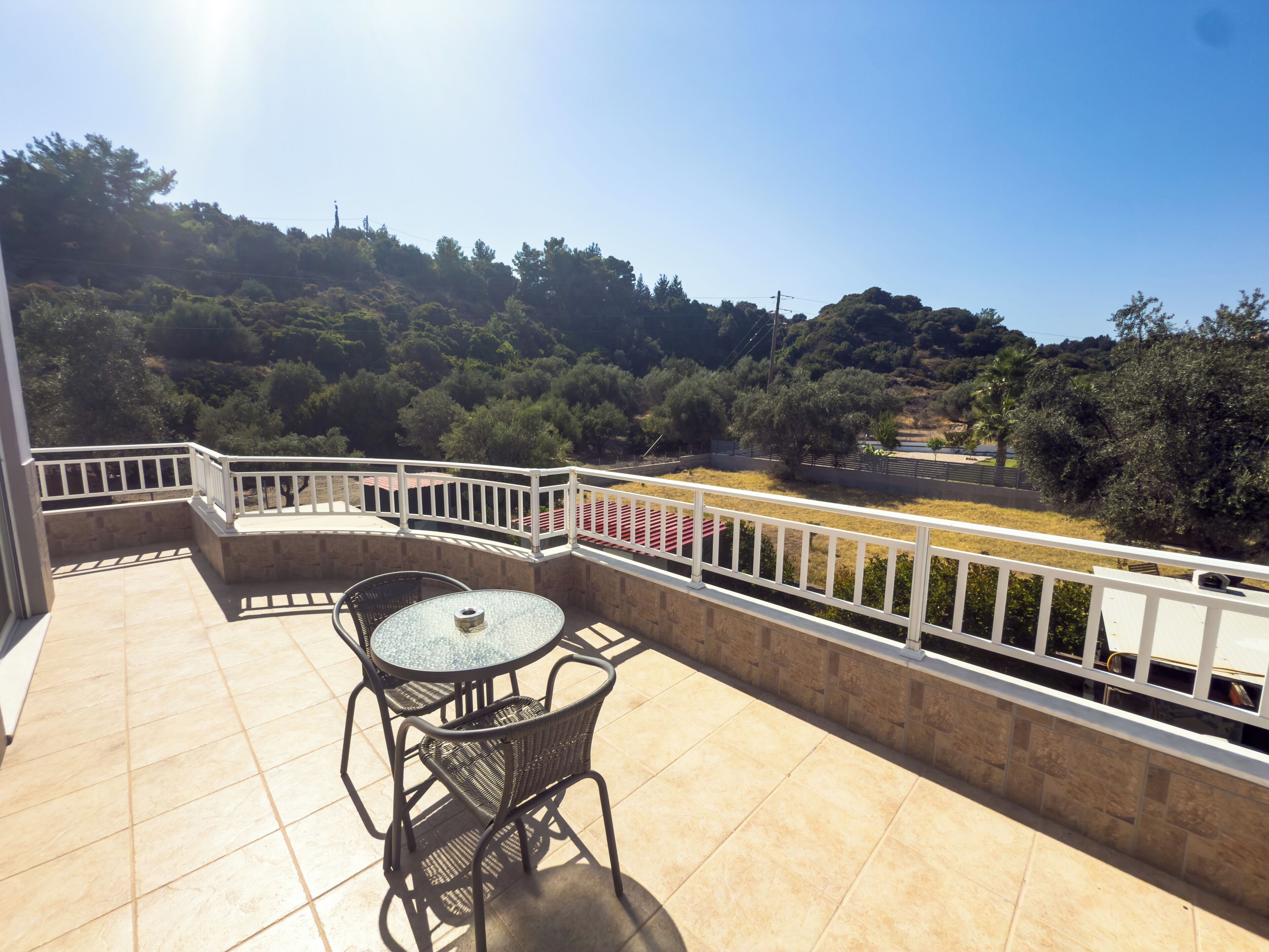 A balcony with a table and chairs, a view of a hilly landscape, and a white railing.