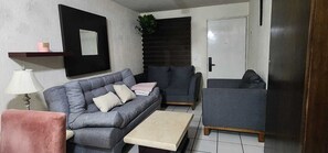 A living room with a grey sofa, a smaller grey armchair, a beige coffee table, a pink chair, a lamp, a mirror, and a door with a plant beside it.