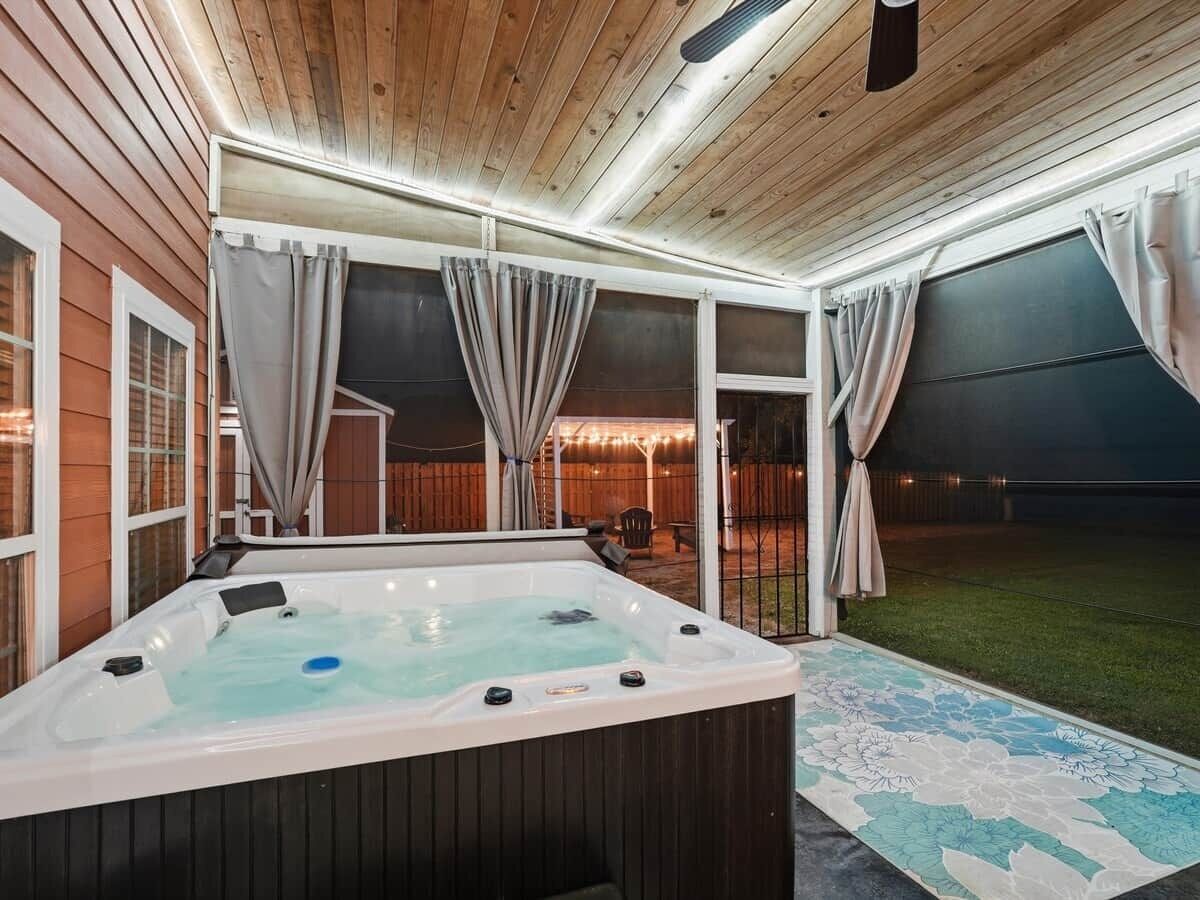 A hot tub with a wooden deck and curtains, surrounded by a wooden fence and a grassy yard.