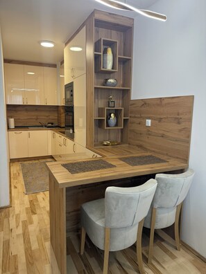 A modern kitchen with wooden cabinetry, a built-in island with bar stools, and a decorative shelf unit. - Daky home near AirPort,tram and City center (Sarajevo)