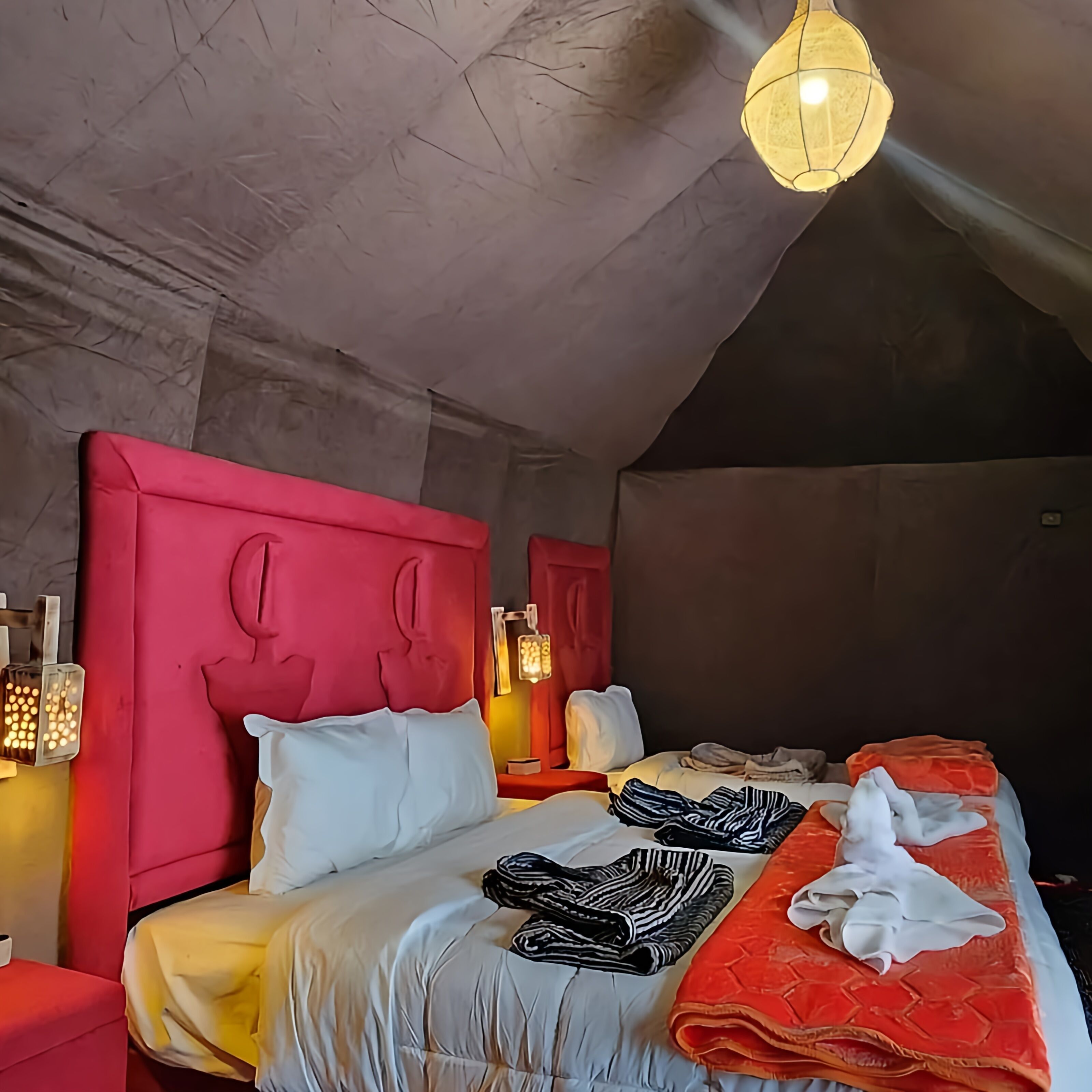 A modern hotel room with a sloped ceiling, a red headboard, a large bed with white and orange bedding, and a hanging light fixture.