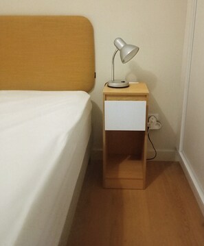 Una cama con sábana blanca, una mesita de noche de madera con una lámpara y un enchufe.