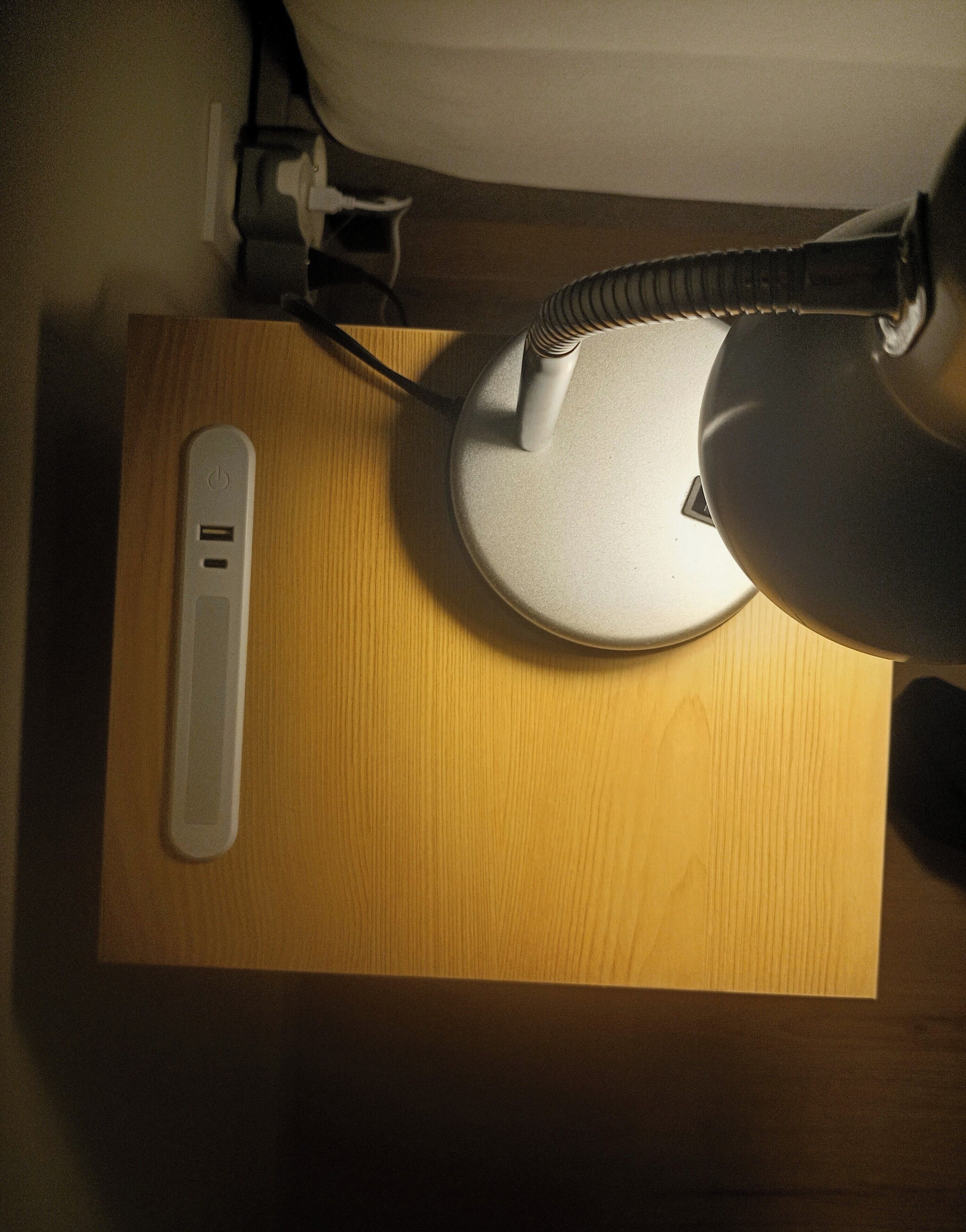 A wooden nightstand with a lamp, a power strip, and a device with a power button.