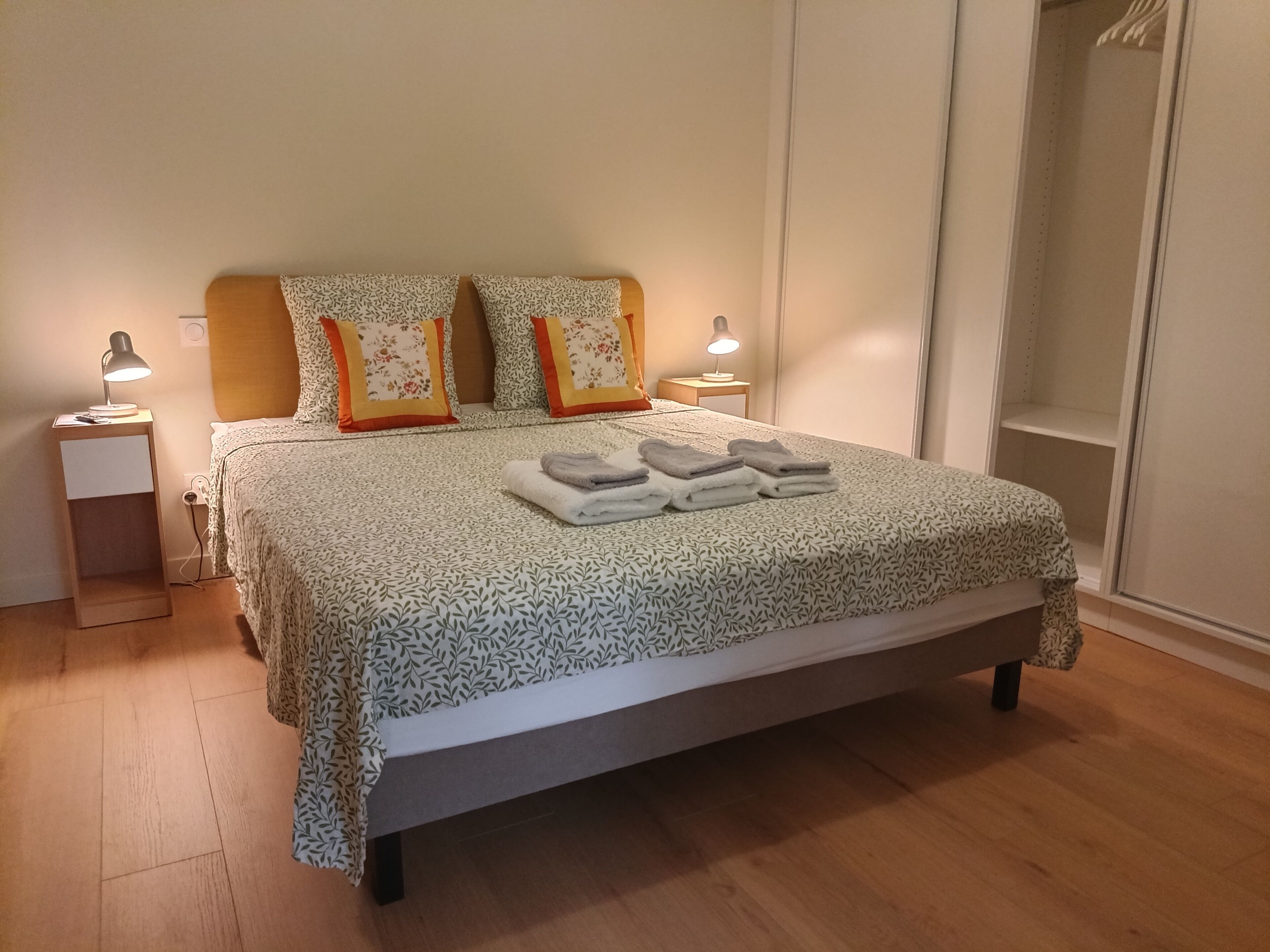 Un lit bien fait, recouvert d’une couvre-lit à motifs, avec deux lampes de chevet et une pile de serviettes pliées.