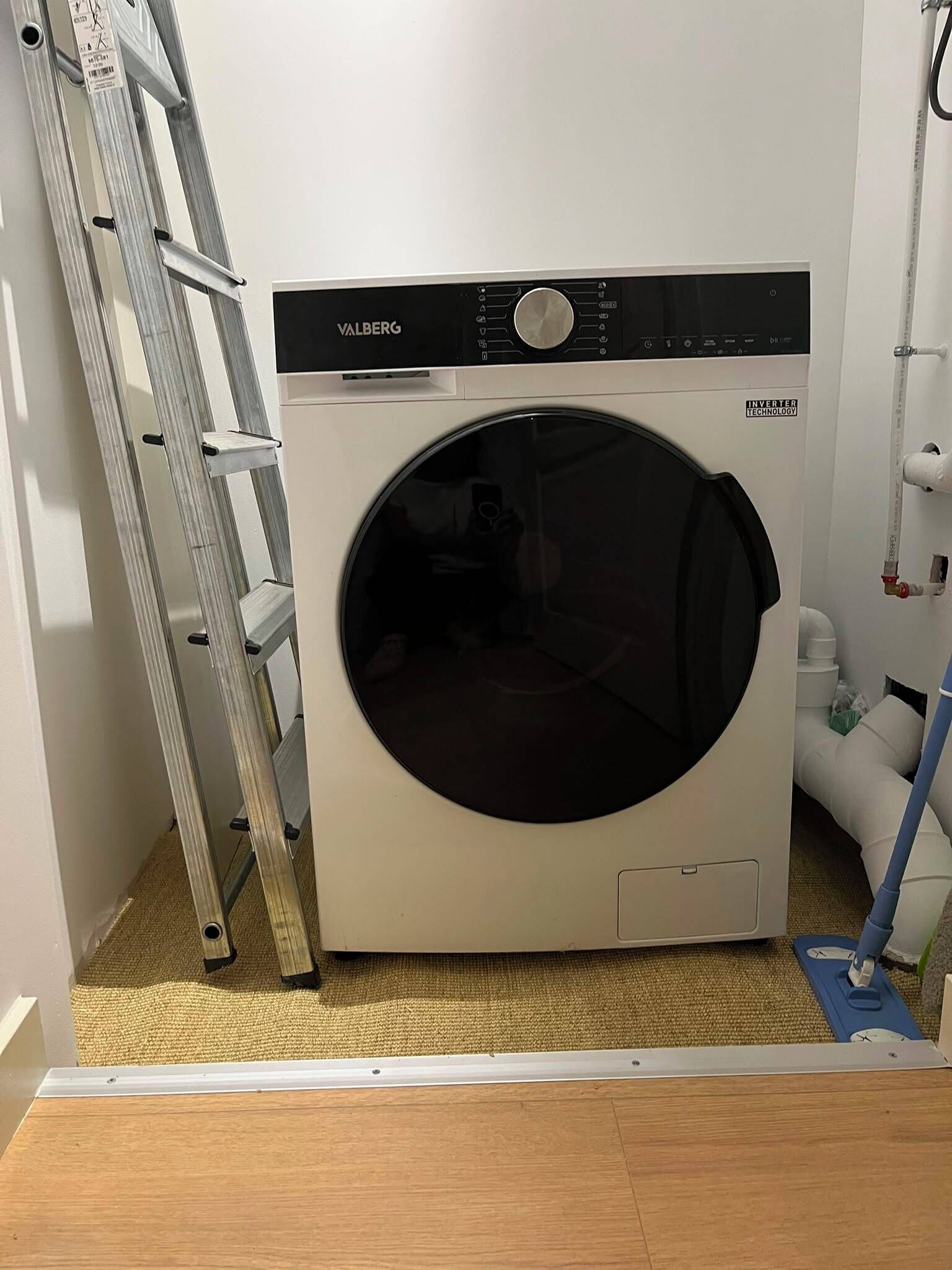 A front-loading washing machine with a control panel, a ladder, and a mop leaning against the wall.