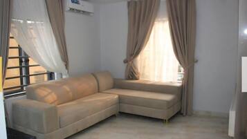 Una sala de estar con un sofá seccional color beige, una ventana con cortinas y un equipo de aire acondicionado.