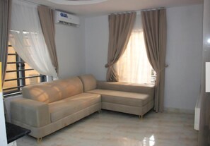 Una sala de estar con un sofá seccional color beige, una ventana con cortinas y un equipo de aire acondicionado.