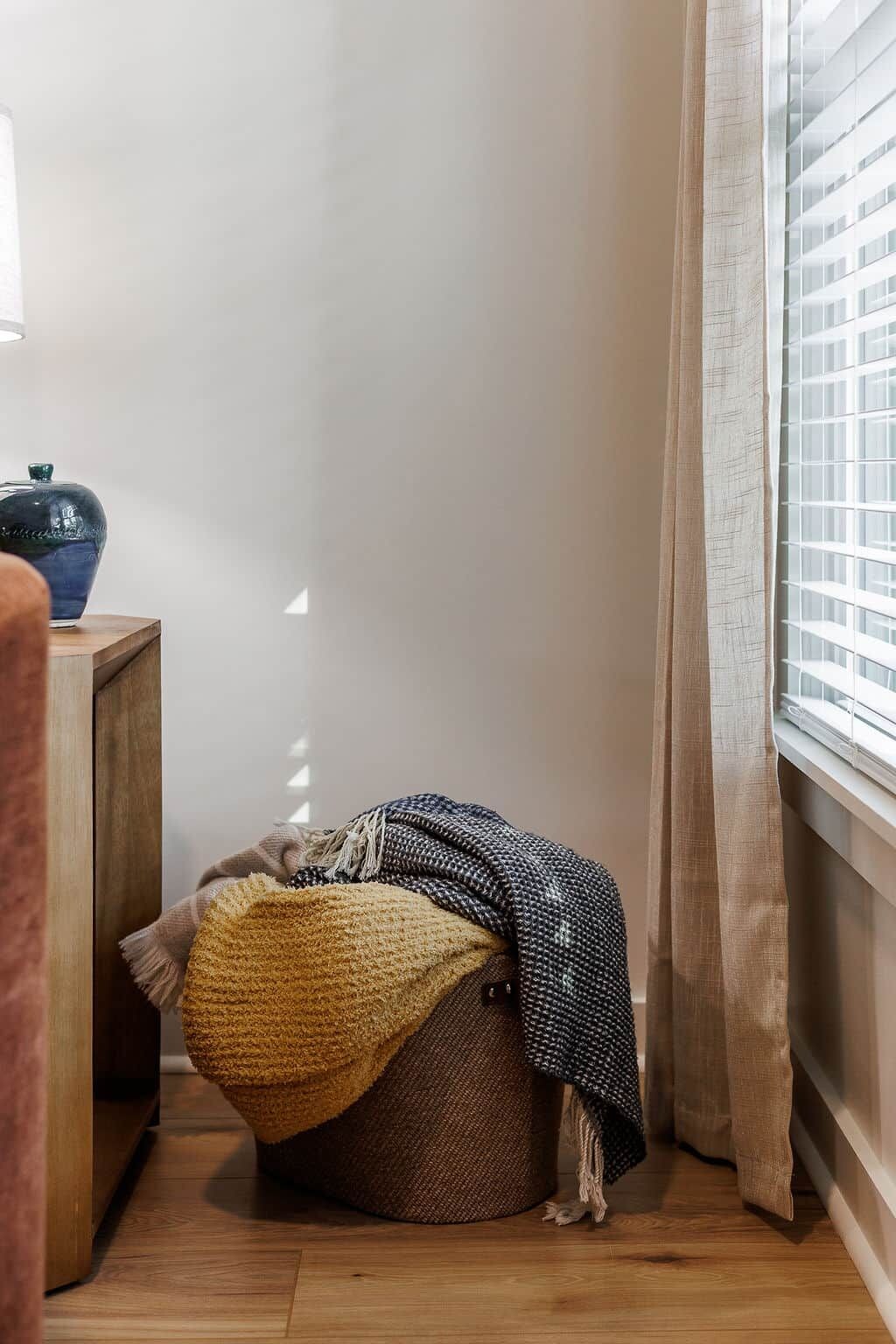 A corner of a room with a wooden floor, a small wooden side table with a blue vase, a bean bag chair with a yellow cushion, and a window with blinds.
