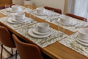 Mesa de comedor dispuesta con platos, tazones y cubiertos blancos, y un camino de mesa de encaje.