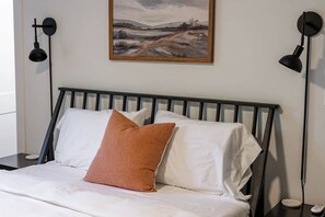Een bed met witte beddengoed en één bruin kussen, een zwart hoofdeinde en een schilderij van een landschap aan de muur.