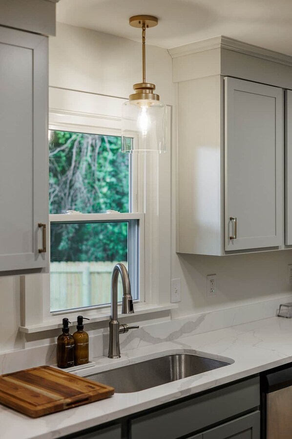 A kitchen with white cabinetry, a stainless steel sink, a wooden cutting board, and a window with a view of greenery.