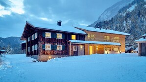 Un chalet en bois doté de nombreuses fenêtres et d’un toit enneigé, entouré d’un paysage hivernal et de montagnes.