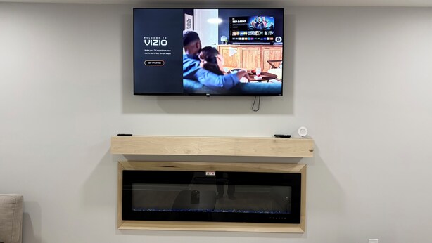 A flat-screen TV mounted on a wall displaying a welcome screen for a service. Below the TV, there is a wooden shelf with a remote control and a white circular object.