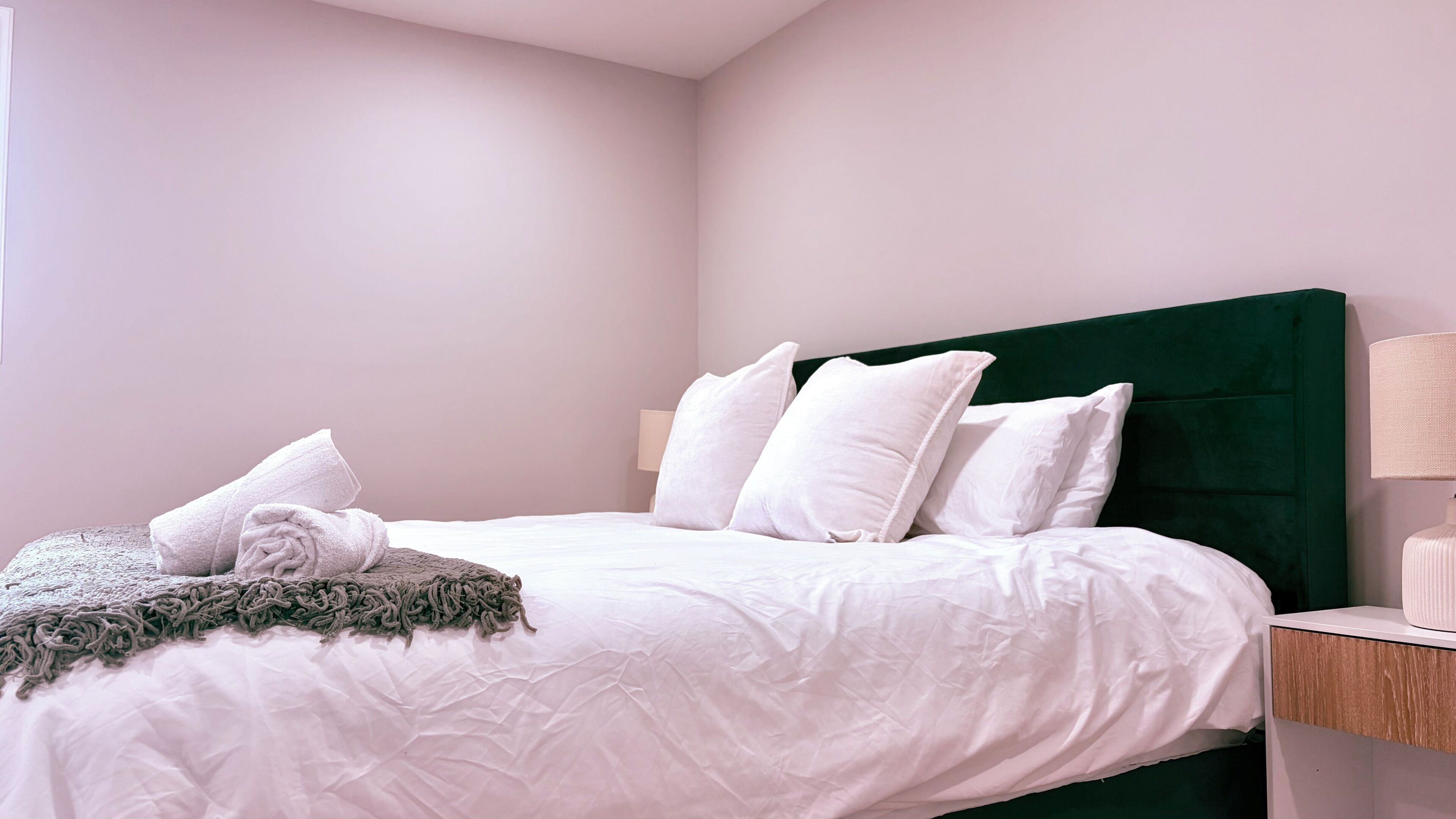 Uma cama bem arrumada com roupa de cama e almofadas brancas, cabeceira verde e mesa de cabeceira com abajur.