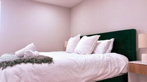 Uma cama bem arrumada com roupa de cama e almofadas brancas, cabeceira verde e mesa de cabeceira com abajur.