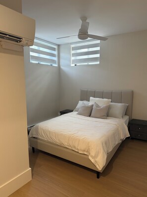 Un dormitorio con cama, mesitas de noche, ventilador de techo y dos ventanas con persianas.