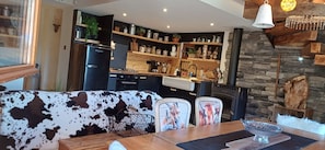 A cozy living room with a cow-print sofa, a dining table with a decorative bowl, and a kitchen with wooden shelves and a stone fireplace. - Alpine chalet in the heart of the Ecrins National Park (L'Argentière-la-Bessée)