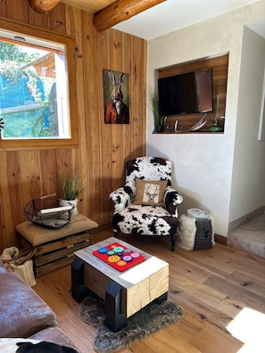 A cozy room with wooden walls, a cow-patterned armchair, a television, and a window with a view of greenery. - Alpine chalet in the heart of the Ecrins National Park (L'Argentière-la-Bessée)
