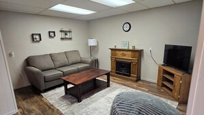 Una sala de estar con sofá, mesa de centro, reposapiés, chimenea, mueble para televisor y un reloj en la pared.