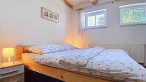A bedroom with a wooden bed, a bedside table with a lamp, a radiator, and a window with a view of trees. - Černý Důl by Interhome (Cerny Dul)