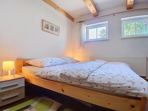 Ein Schlafzimmer mit einem Holzbett, einem Nachttisch mit Lampe, einem Heizkörper und einem Fenster mit Blick auf Bäume.