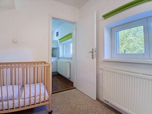 A room with a crib, a window with a view of greenery, and a door leading to a kitchen area. - Černý Důl by Interhome (Cerny Dul)