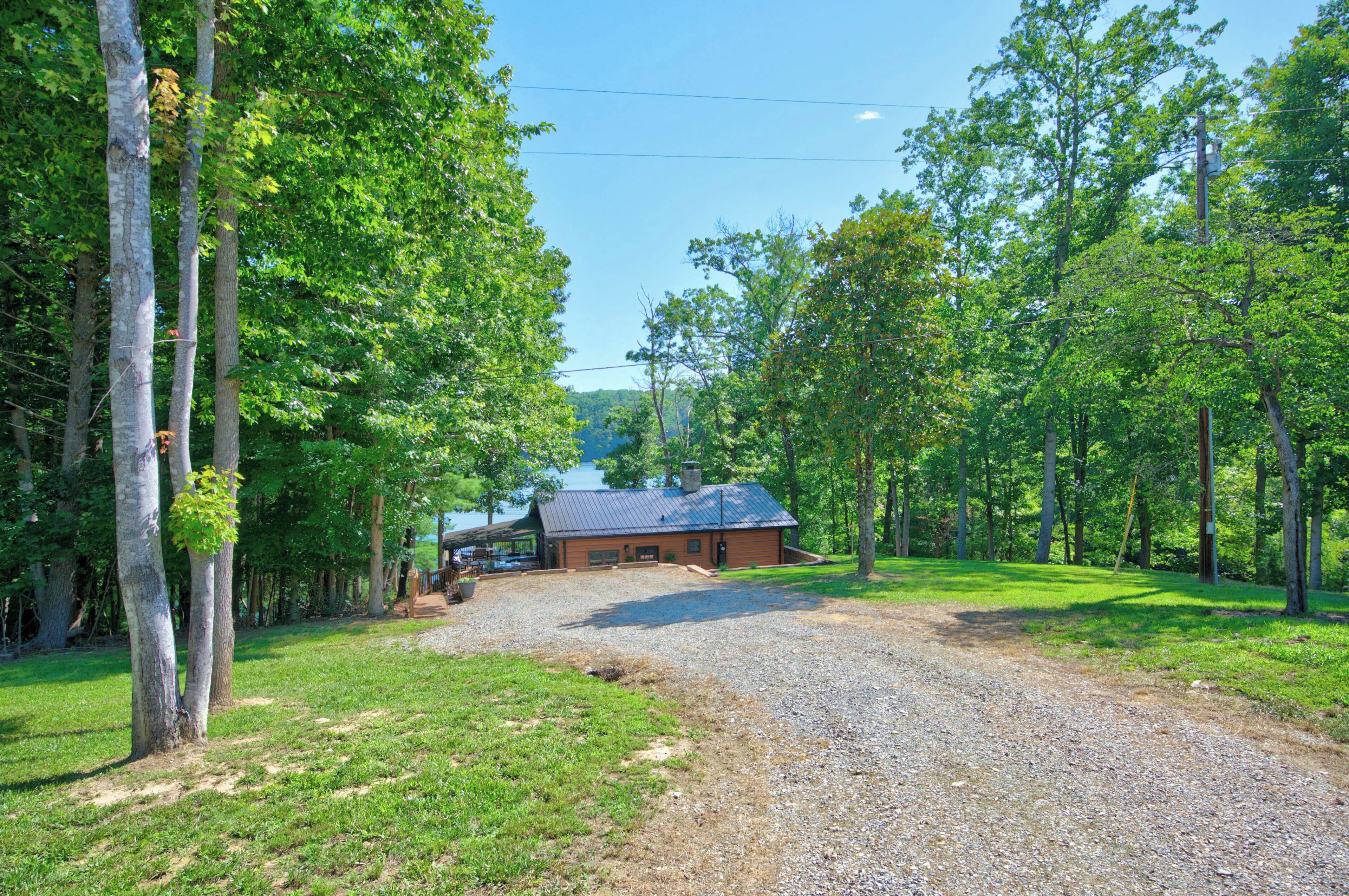 A rustic cabin with a gravel driveway, surrounded by dense trees and a clear sky.