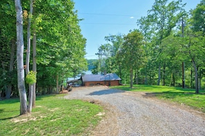 A rustic cabin with a gravel driveway, surrounded by dense trees and a clear sky.
