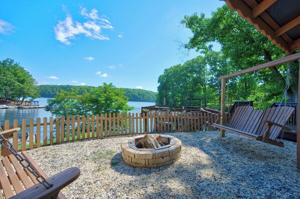 A wooden deck with Adirondack chairs, a fire pit, and a view of a lake and forest.