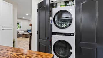 A laundry room with a front-loading washer and dryer, a wooden table in the foreground, and a living area visible through an open door.