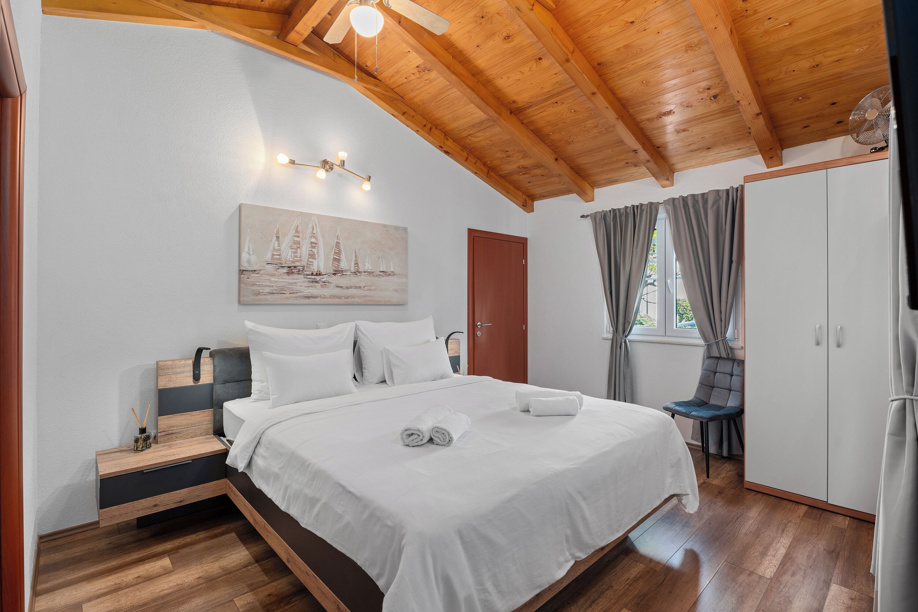 Une chambre avec un plafond en bois, un lit avec une literie blanche, un tableau au mur et une fenêtre avec des rideaux.