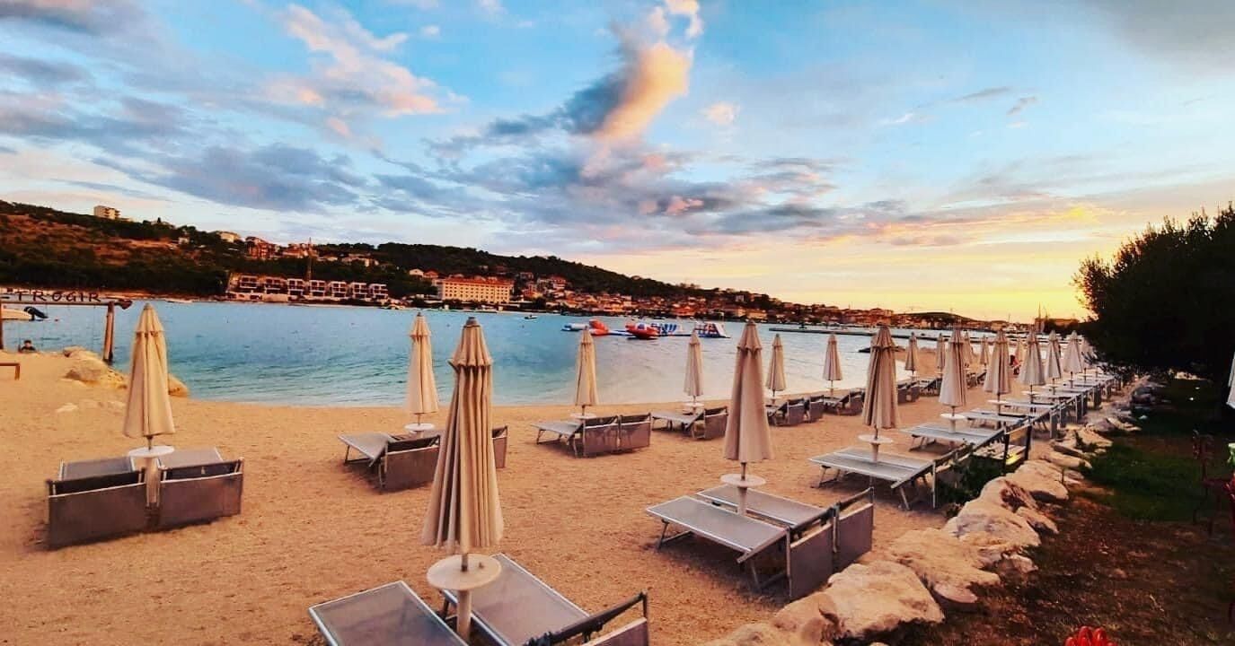 A beach with sun loungers and umbrellas, a clear sky, and a view of the sea and buildings in the distance.