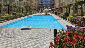 A swimming pool surrounded by multi-story buildings with balconies, a clear sky, and some greenery. - Klara Florenza (Hurghada)
