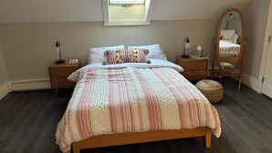 Un dormitorio con tragaluz, cama con ropa de cama estampada, mesita de noche con lámpara, espejo y una alfombra redonda pequeña.