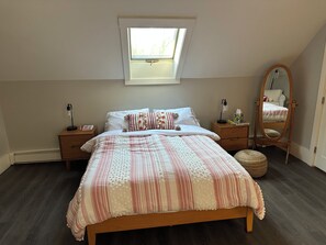 Ein Schlafzimmer mit Dachfenster, einem Bett mit gemusterten Betttextilien, einem Nachttisch mit Lampe, einem Spiegel und einem kleinen runden Teppich.