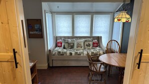 Una habitación acogedora con suelo de madera, una mesa de comedor con sillas, un banco acolchado con cojines estampados y una ventana con persianas.