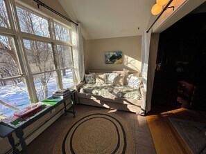 Ein gemütliches Zimmer mit einem großen Fenster, einem Sofa, einem kleinen Tisch mit Büchern und einem Bild an der Wand.