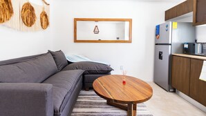 En moderne stue med en grå sofa, et træsofabord og et køkkenområde med et køleskab og en mikrobølgeovn.