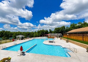 A large outdoor swimming pool with multiple lanes, surrounded by lounge chairs and umbrellas, with a wooden fence and a building in the background. - Pocono Pines Retreat Hot Tub & Cozy Escape in PCP (Tobyhanna)